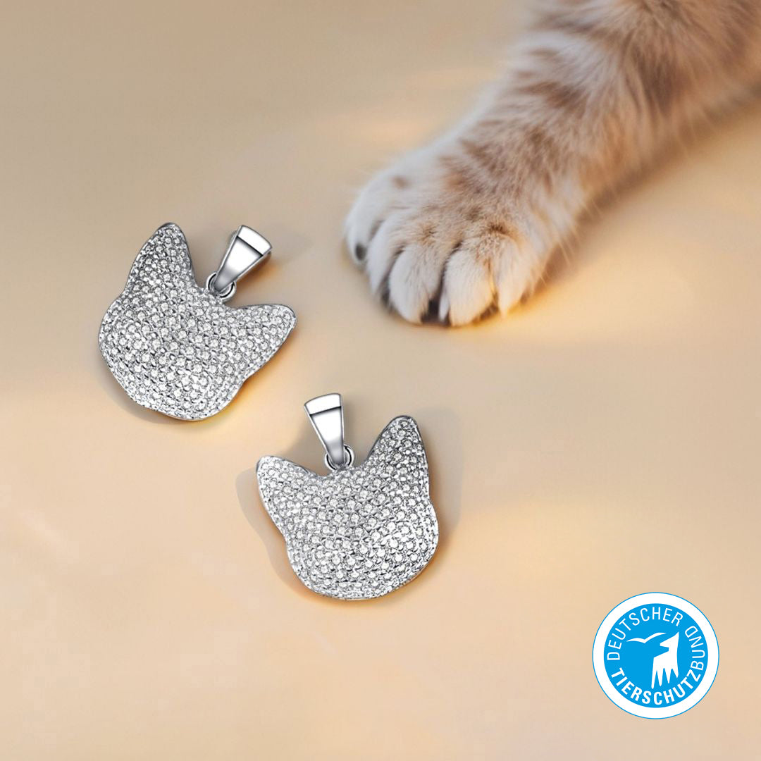 Two silver Shiro cat-head-shaped pendants encrusted with sparkling cubic zirconia, placed beside a soft, outstretched cat paw on a beige background, featuring the Deutscher Tierschutzbund logo in the corner.