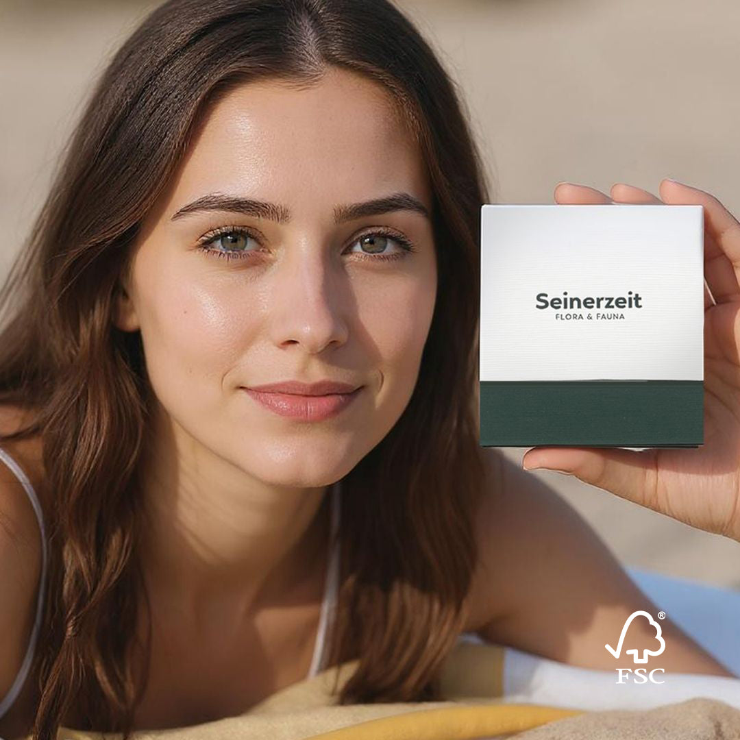Une jeune femme aux longs cheveux bruns et au maquillage naturel sourit doucement en tenant une boîte à bijoux Seinerzeit Flora & Fauna, avec un emballage vert et blanc et le logo de durabilité FSC dans le coin.