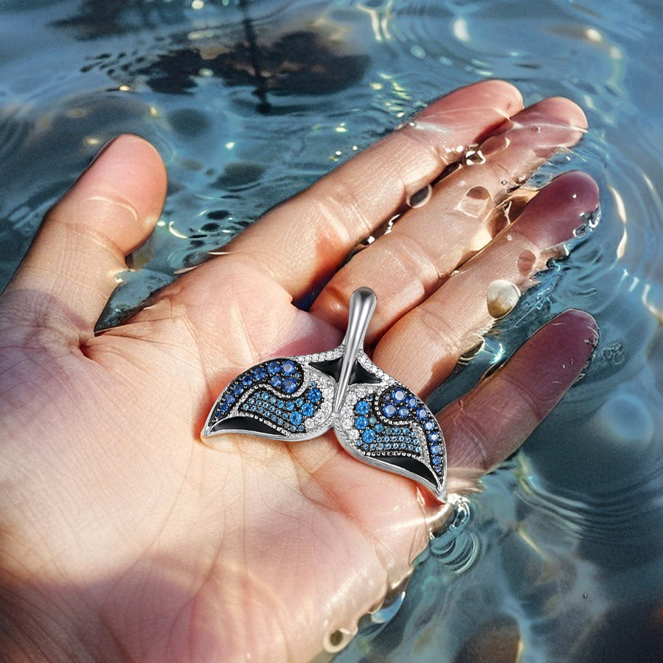 Nessie Fin pendant by Seinerzeit – ocean-inspired sterling silver jewelry with blue and white CZ stones, shaped like a whale tail and photographed in hand in the sea

