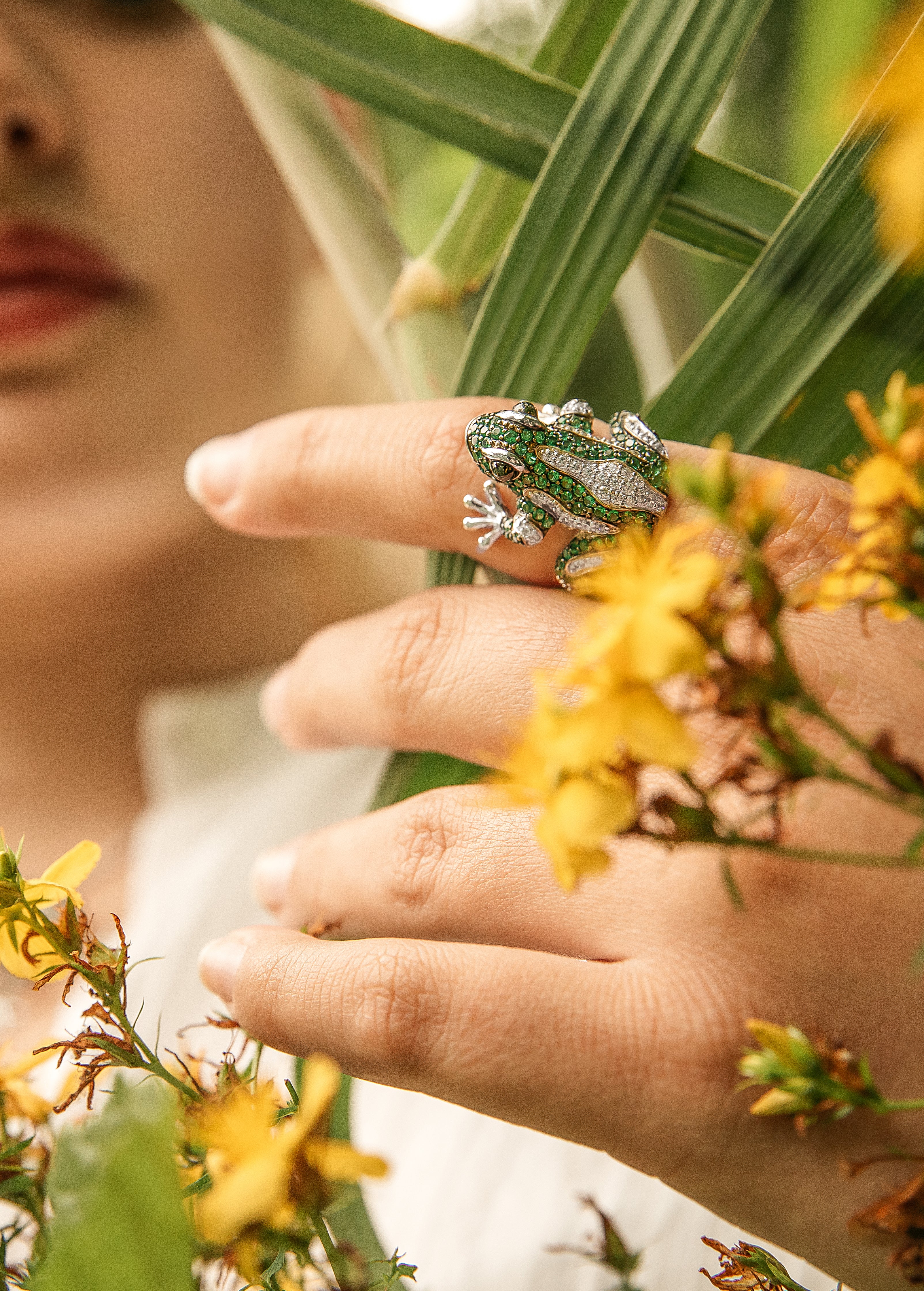 Close-up of a hand wearing the Allen Frog ring, designed with green and silver details, surrounded by yellow flowers and greenery.