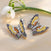 Grace butterfly pendants in sterling silver, adorned with multicolored cubic zirconia stones in yellow, orange, green, purple, and pink, placed beside a white snowdrop flower.
