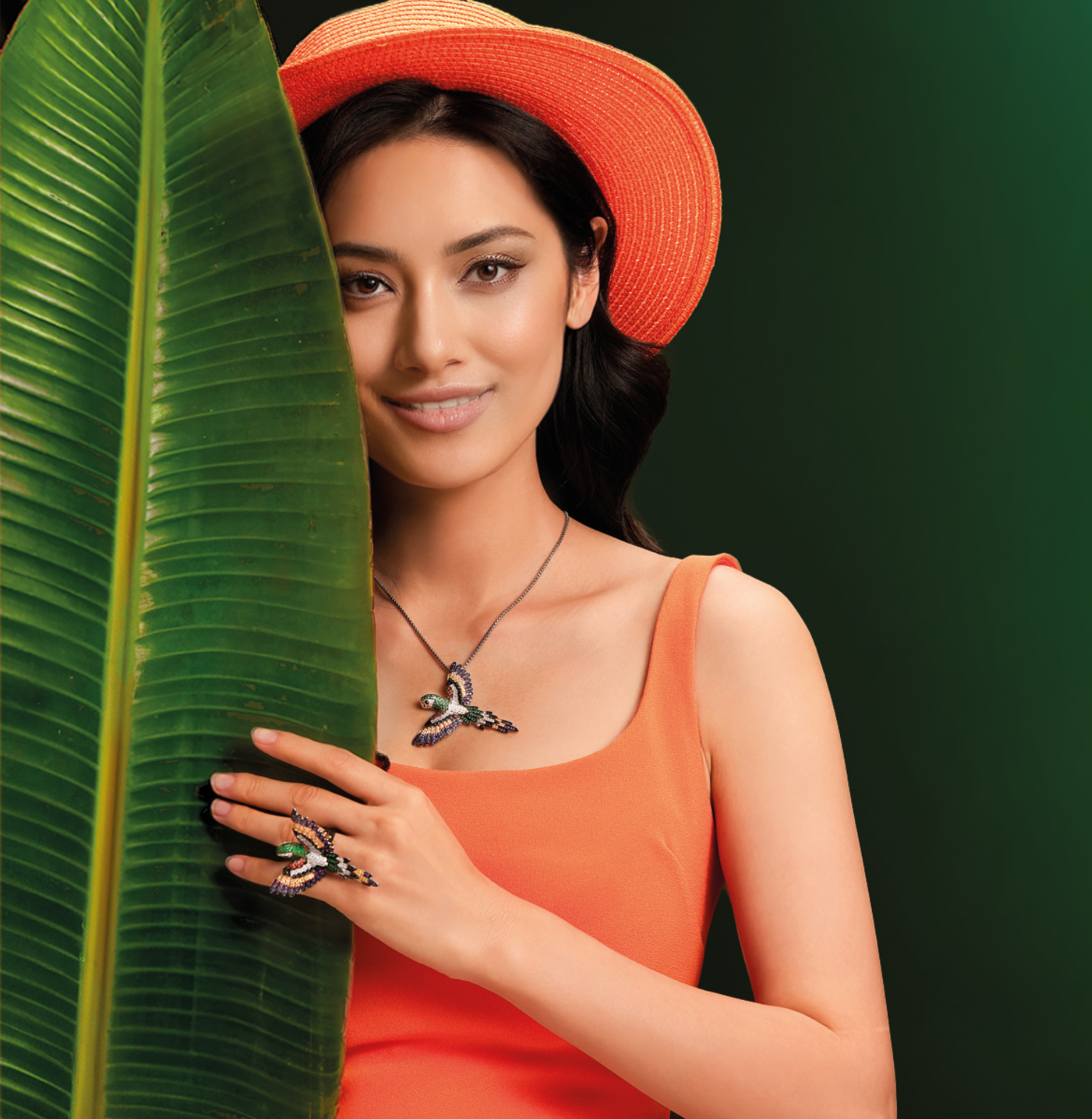Woman in an orange dress and matching hat poses with a large tropical leaf, wearing Seinerzeit Flora & Fauna silver jewelry set featuring a colorful parrot pendant and matching ring from the Antoine Papagei Jungle Adventure collection.