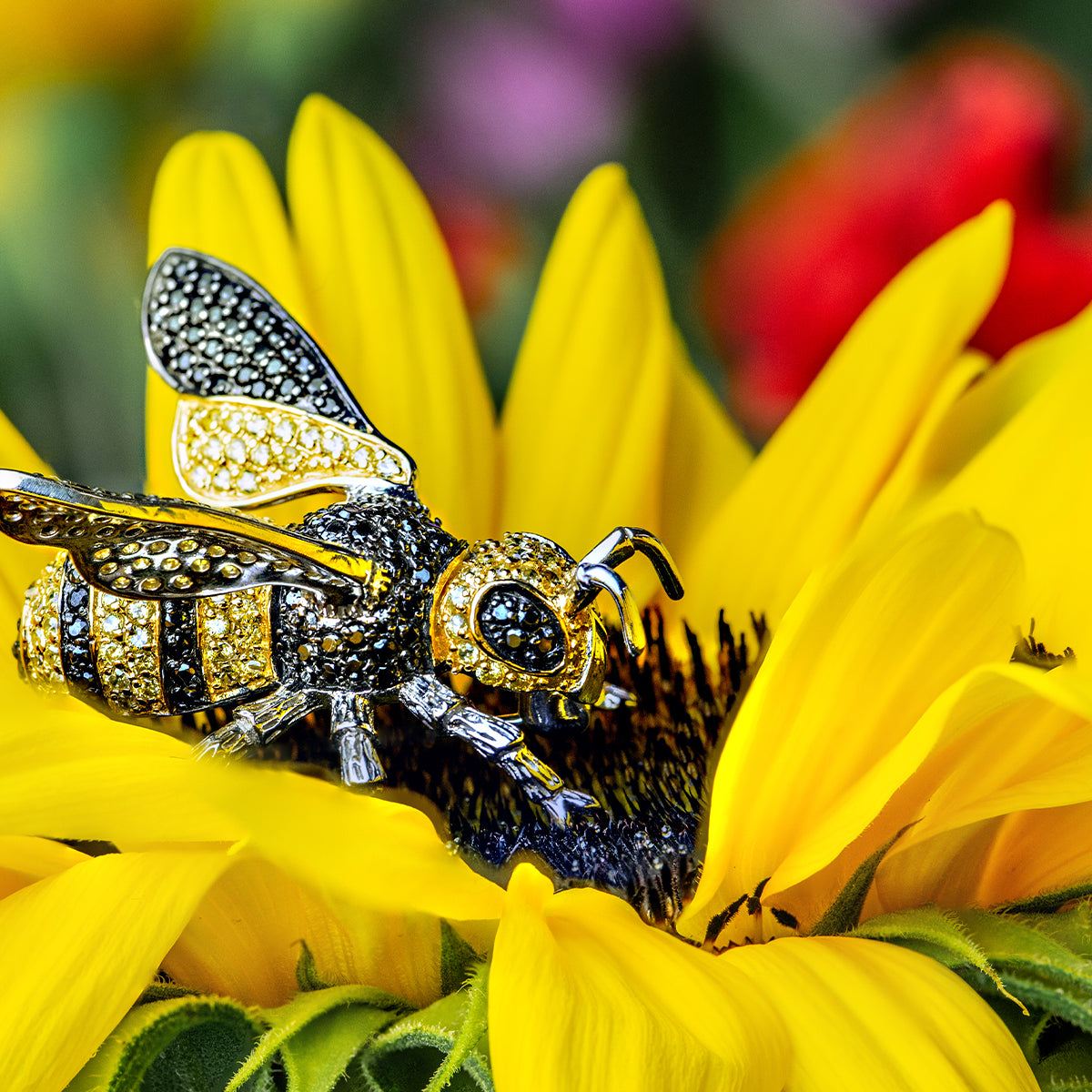 Close-up of the Seinerzeit Flora & Fauna Vesna bee pendant, adorned with yellow and black cubic zirconia, resting on the center of a vibrant yellow sunflower with blurred colorful flowers in the background.