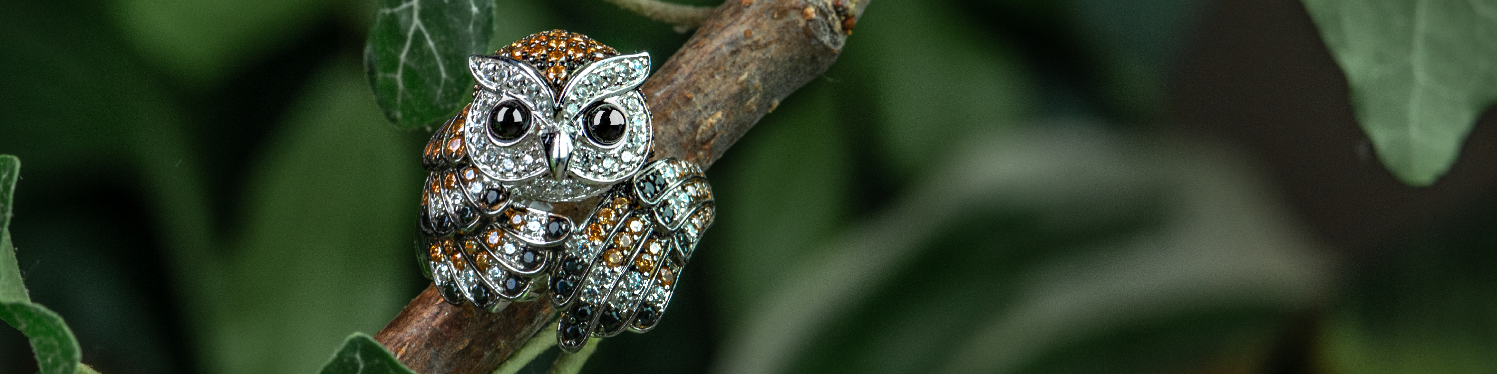 Close-up of the Alex Owl Ring, designed in sterling silver with intricate detailing. The ring features an owl perched on a branch, adorned with sparkling stones in shades of amber, white, and green. The background is blurred green foliage, emphasizing the natural, woodland-inspired theme of the jewelry piece.