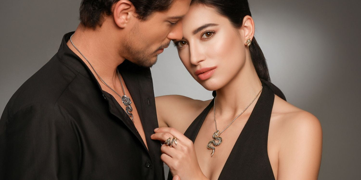Elegant couple wearing matching dragon-themed jewelry from Seinerzeit Flora & Fauna — the woman styled in the Belarius snake pendant, Drakonis dragon stud earrings, and a statement ring, while the man wears the Drakonis dragon pendant — both dressed in black against a dark studio background