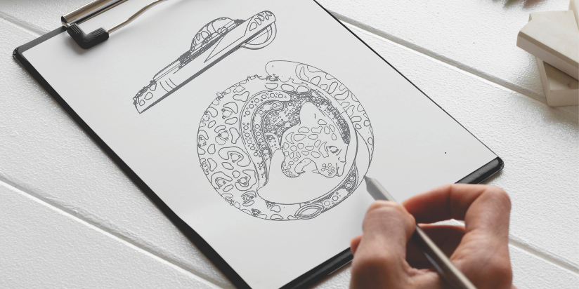 Close-up of a hand sketching a detailed leopard pendant design on a clipboard, surrounded by colored pencils and art supplies on a white wooden table.