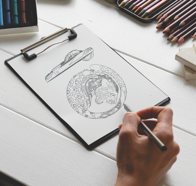 Close-up of a hand sketching a detailed leopard pendant design on a clipboard, surrounded by colored pencils and art supplies on a white wooden table.