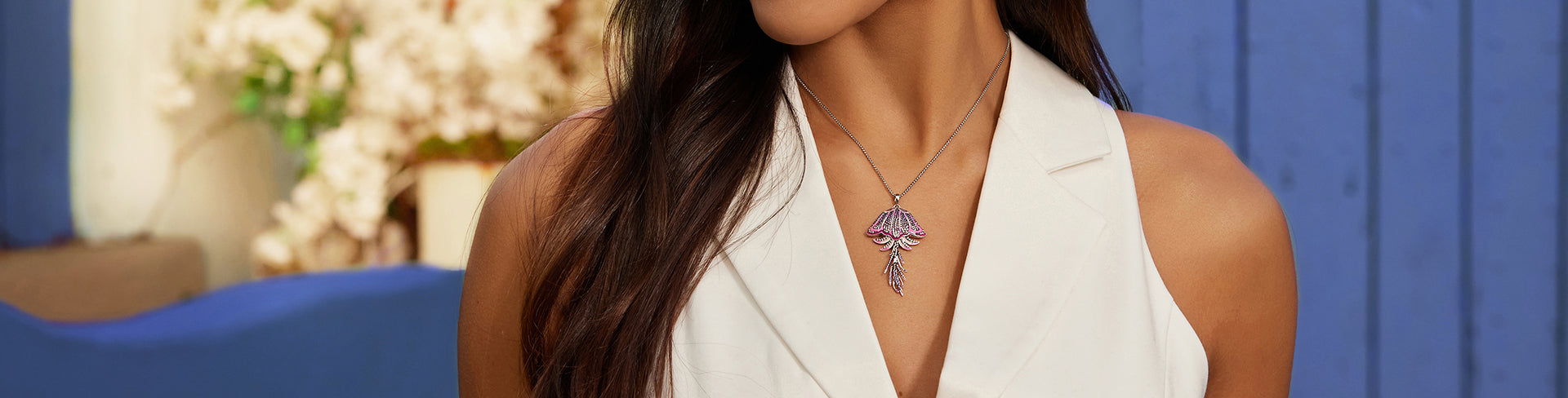 Banner showing the Trixie Jellyfish pendant in sterling silver with pink and purple cubic zirconia stones. The pendant is worn on a silver chain by a woman dressed in a sleeveless white blazer, set against a background of blurred flowers and a vivid blue wooden wall.